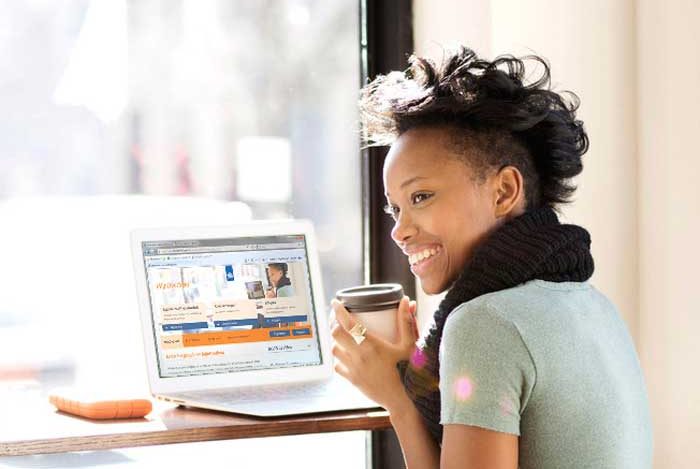Vrolijke jonge mevrouw met kop koffie in haar handen achter een laptop